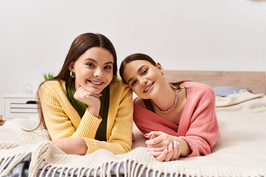 Dos mujeres jóvenes con atuendo casual, acostadas en una cama, sonriendo calurosamente a la cámara en un momento acogedor e íntimo en casa. - Foto, imagen