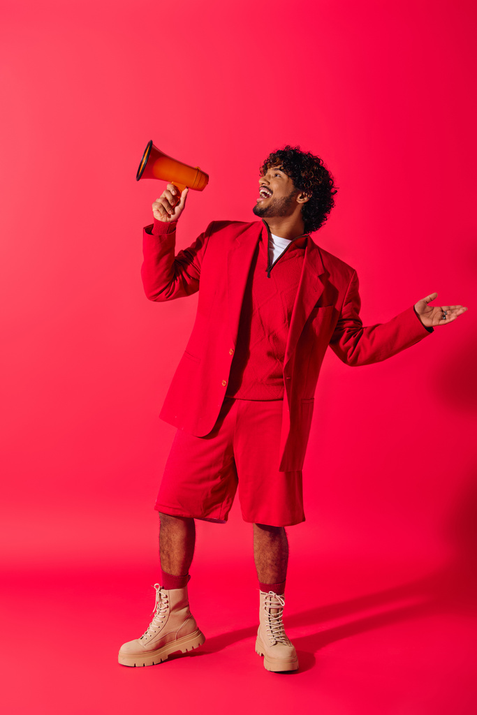 Canlı kırmızı takım elbiseli genç Hintli adam canlı bir arkaplana karşı kırmızı bir megafon tutuyor.. - Fotoğraf, Görsel