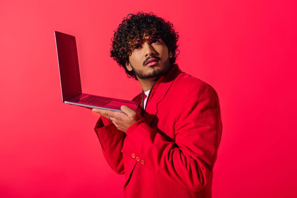 Przystojny młody Indianin w czerwonym garniturze pewnie trzyma laptopa. - Zdjęcie, obraz