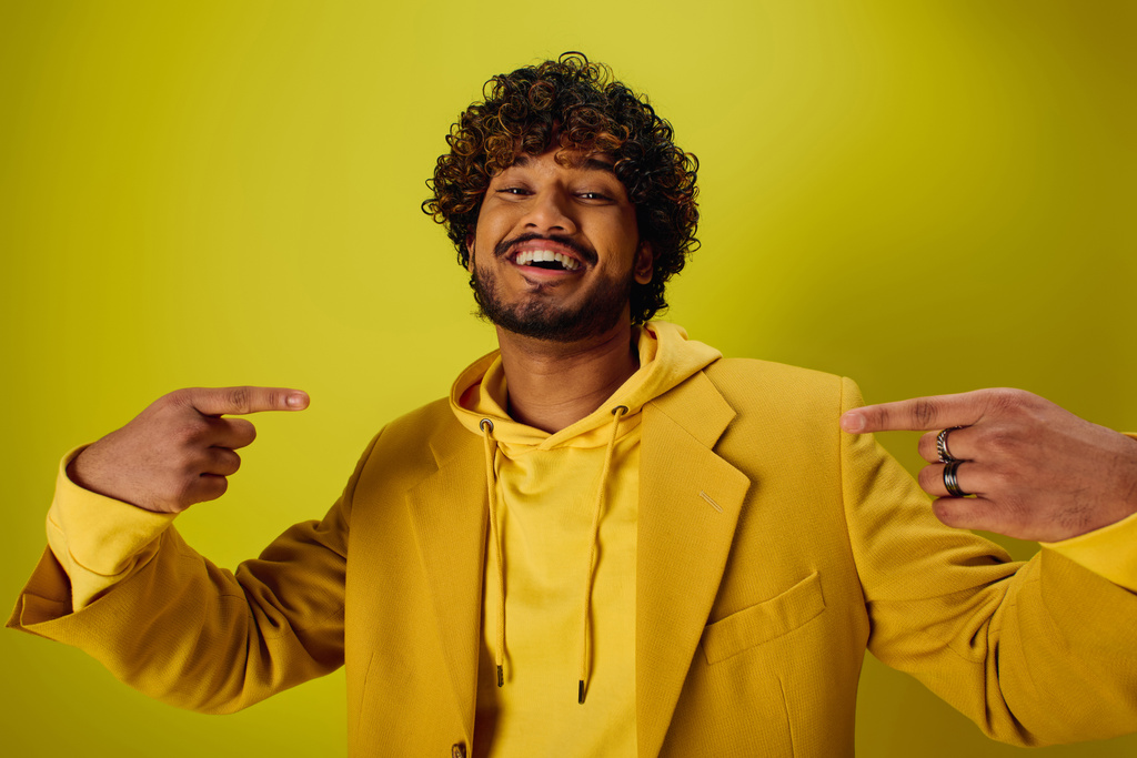 Ein gutaussehender junger indischer Mann in einer gelben Jacke zeigt auf etwas vor einer lebhaften Kulisse. - Foto, Bild
