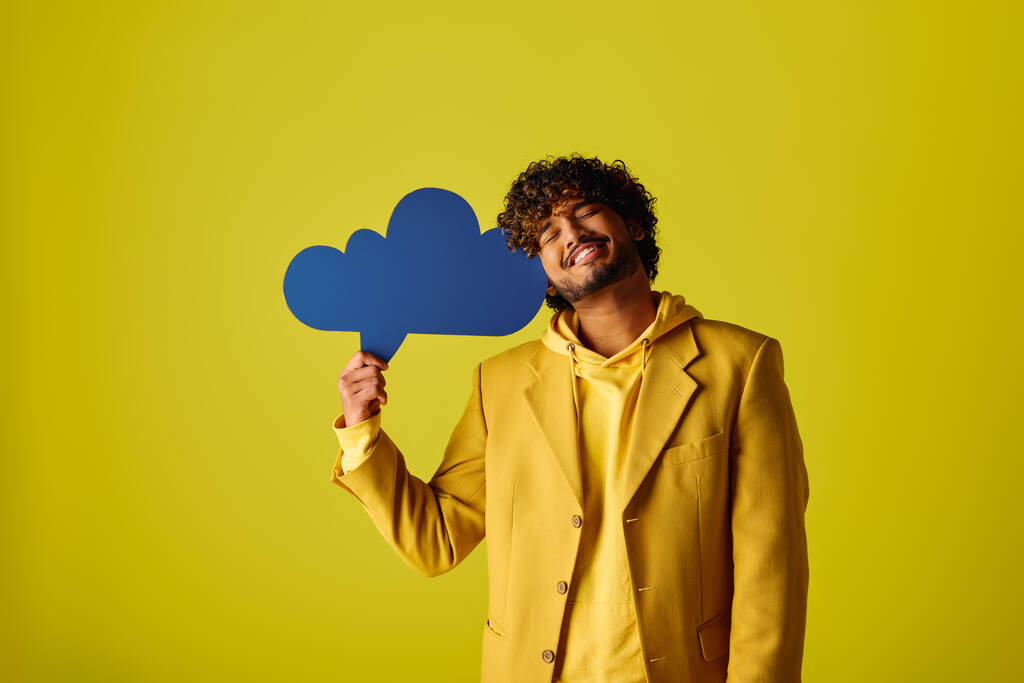 Handsome young Indian man in yellow suit holding blue speech bubble on vivid backdrop. - Photo, Image