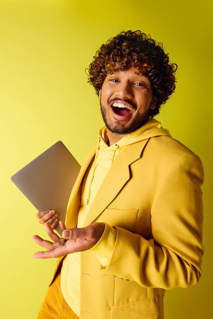 Hombre indio guapo en un traje amarillo vibrante con confianza sostiene una computadora portátil. - Foto, imagen