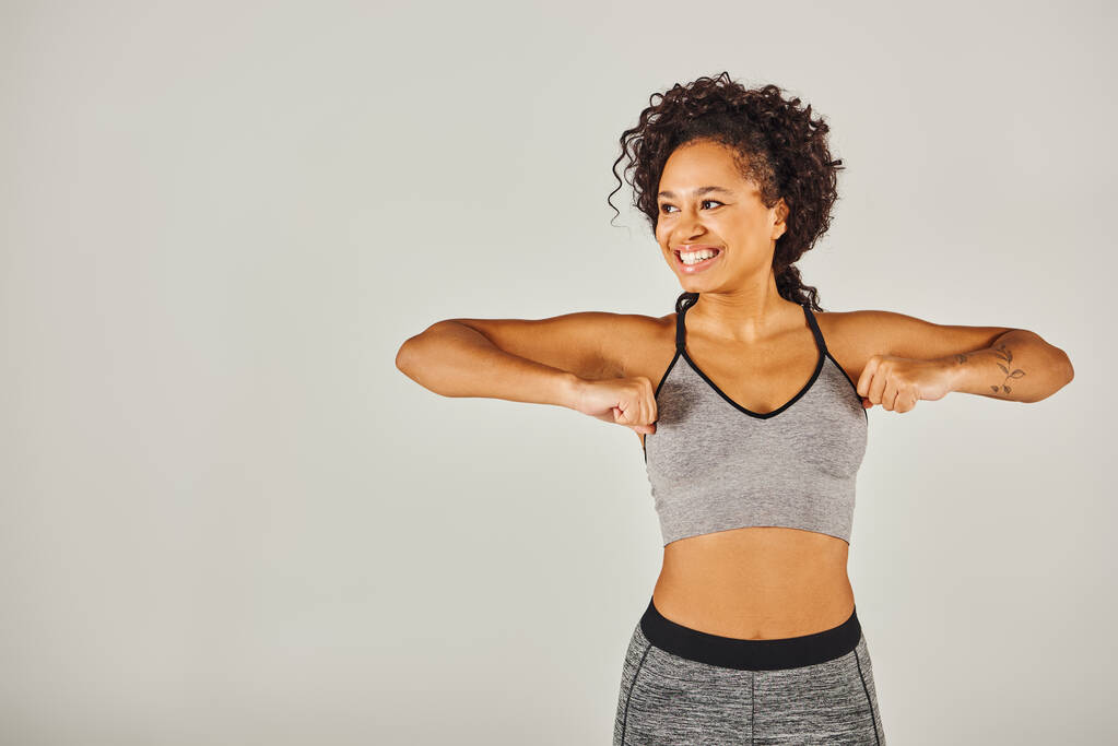 Una giovane donna afroamericana in activewear energicamente flettendo le braccia davanti a uno sfondo grigio. - Foto, immagini