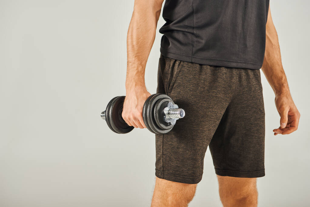 A young sportsman in active wear energetically lifts a pair of dumbbells in a studio with a grey background. - Photo, Image