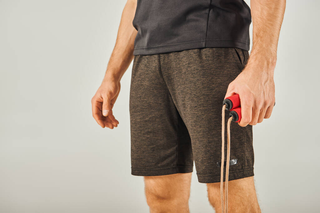 Молодий спортсмен в активному одязі вміло тримає яскраву червоно-чорну мотузку в студії з сірим фоном. - Фото, зображення
