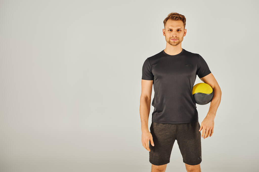 Jovem desportista de t-shirt preta demonstrando exercício físico com uma bola amarela e preta vibrante em estúdio. - Foto, Imagem
