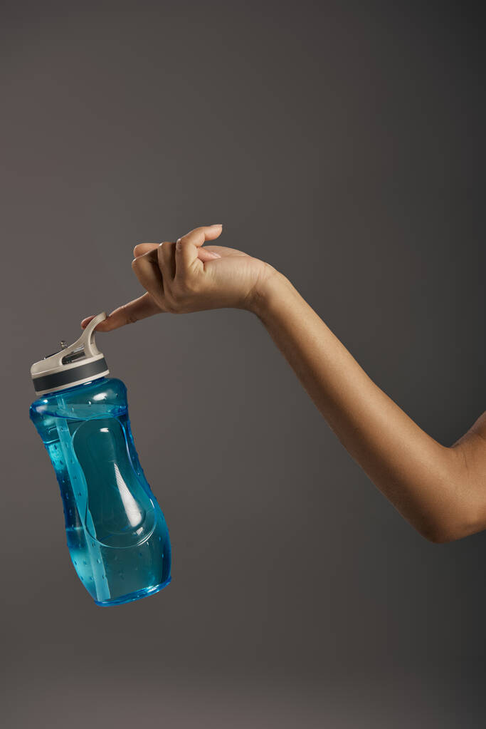 Amerikalı sporcu kadın, gri bir arka planda mavi bir su şişesi tutuyor.. - Fotoğraf, Görsel