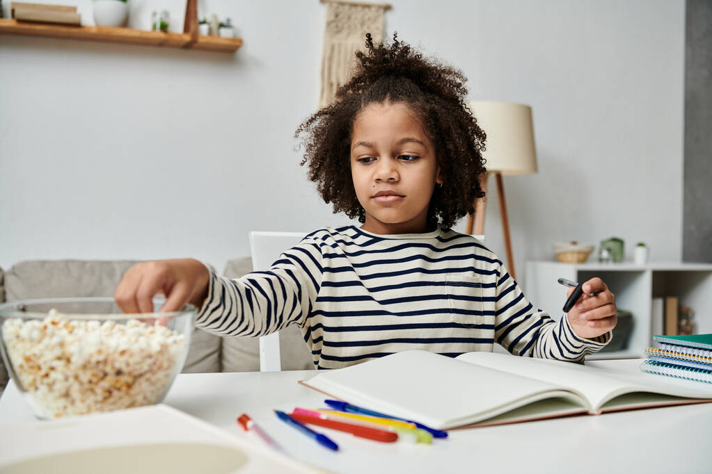 Ένα μικρό κορίτσι κάθεται σε ένα τραπέζι με ένα μπολ ποπ κορν και ένα βιβλίο, απολαμβάνοντας μια στιγμή ηρεμίας και φαντασίας - Φωτογραφία, εικόνα