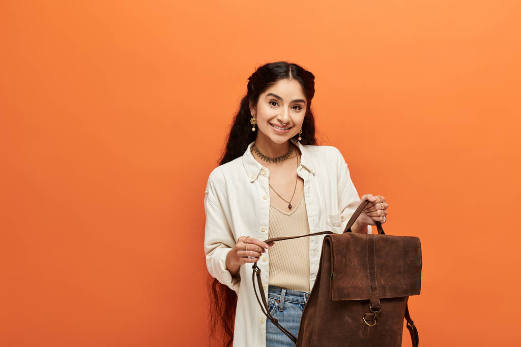 Young indian woman holding backpack against vibrant orange background. - Photo, Image