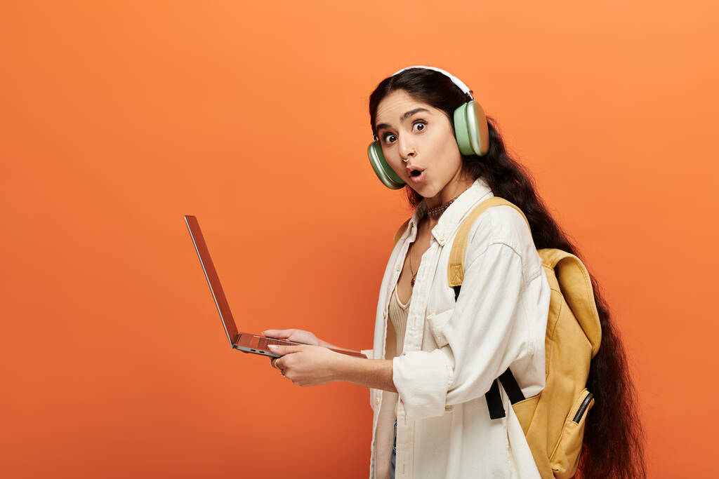Młoda Hinduska kobieta ze słuchawkami, korzystająca z laptopa na pomarańczowym tle. - Zdjęcie, obraz