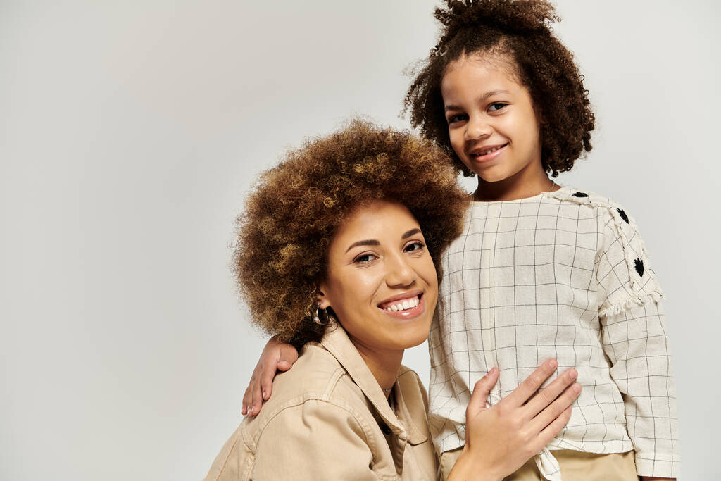 Lockige afroamerikanische Mutter und Tochter in modischer Kleidung posieren gemeinsam vor schickem grauen Hintergrund. - Foto, Bild