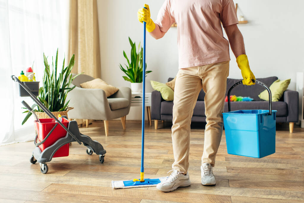 Hezký muž v útulných domácích šatech pečlivě čistí podlahy s kýblem čistících prostředků. - Fotografie, Obrázek
