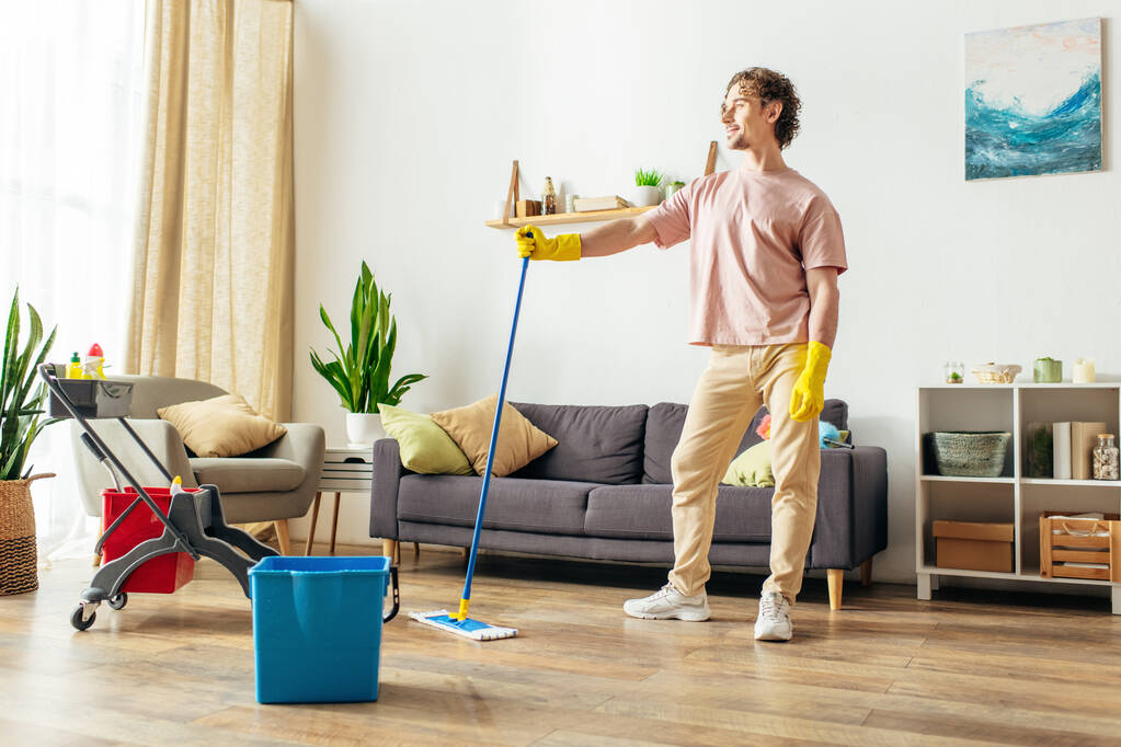 Ein gutaussehender Mann in kuscheliger Hauskleidung putzt das Wohnzimmer mit Wischmopp und Eimer. - Foto, Bild