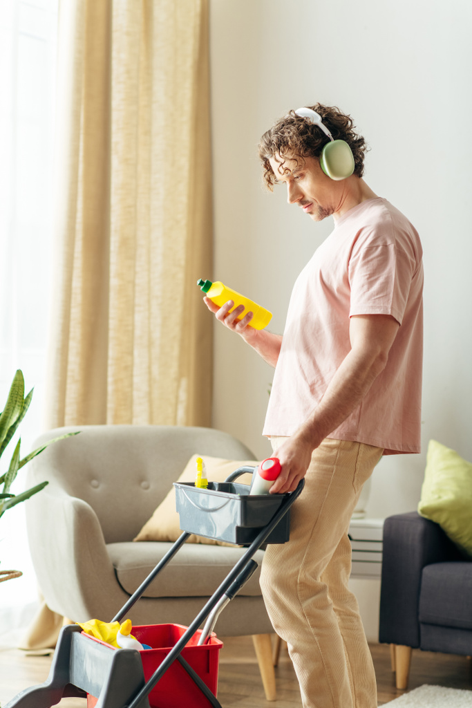 Een stijlvolle man in huiskleding reinigt een gezellige woonkamer met koptelefoon op. - Foto, afbeelding