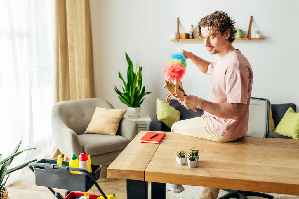 Jóképű férfi kényelmes otthonban játszik egy játékkal, miközben az asztalon takarít.. - Fotó, kép