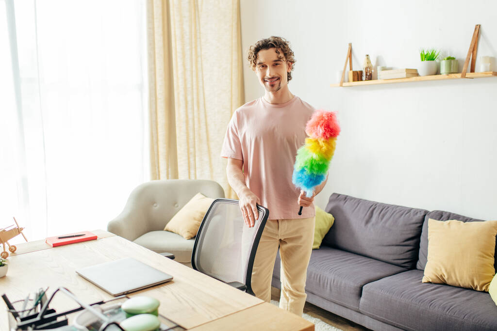 Handsome man in cozy homewear holding a vibrant duster in stylish living room. - Photo, Image