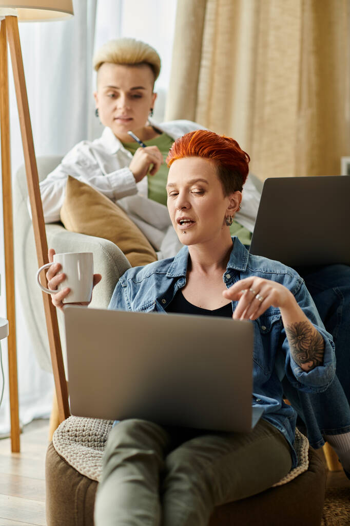 Lesbijska para z krótkimi włosami wygodnie siedzi na kanapie, pochłonięta używaniem laptopa razem w domu. - Zdjęcie, obraz