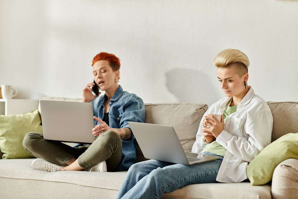 Duas mulheres com cabelo curto sentam-se lado a lado em um sofá, cada uma absorvida em sua própria tela de laptop. - Foto, Imagem