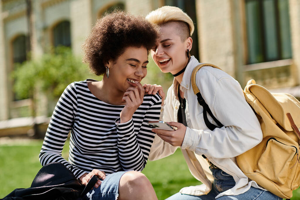 Дві молоді дівчата в стильному вбранні сидять на траві, занурені в телефон, серед безтурботної обстановки. - Фото, зображення