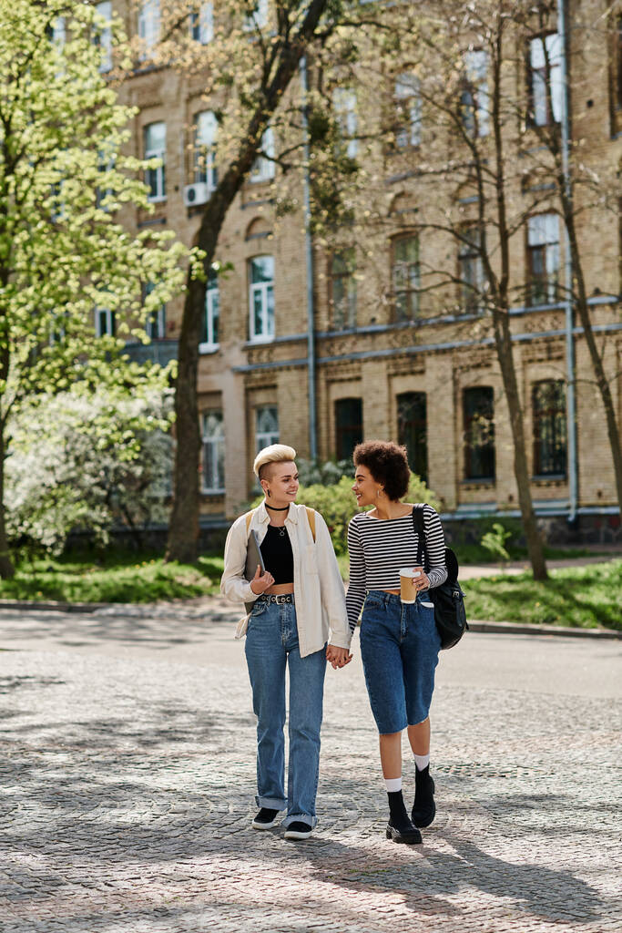 Dos mujeres jóvenes, una pareja lesbiana multicultural, paseando por una calle de la ciudad cerca de un campus universitario con un atuendo elegante. - Foto, Imagen