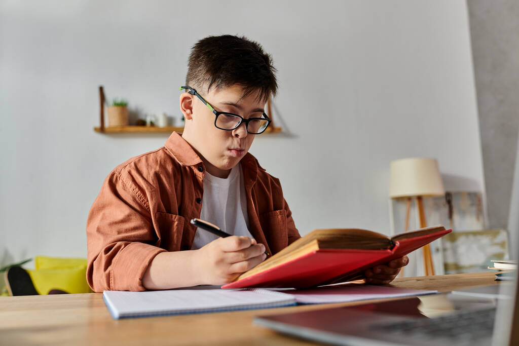 Un niño con síndrome de Down lee un libro y trabaja en un portátil en un escritorio. - Foto, Imagen