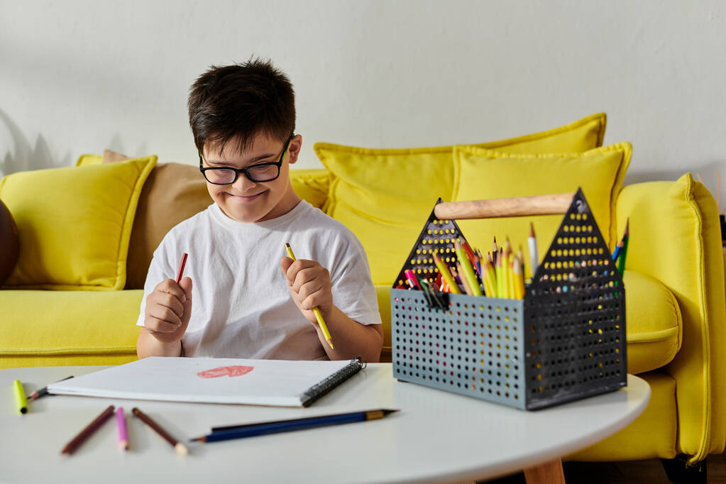 uroczy chłopiec z zespołem Downa siedzi przy stole z kolorowymi ołówkami. - Zdjęcie, obraz