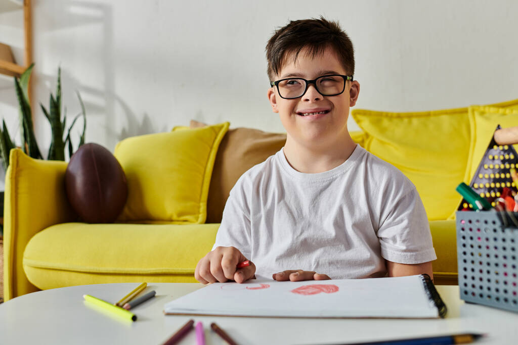 Un adorabile ragazzo con la sindrome di Down con gli occhiali siede a un tavolo, circondato da matite colorate. - Foto, immagini
