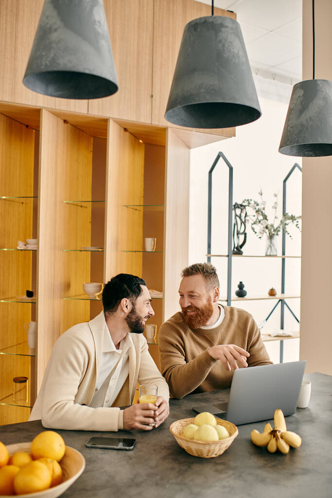 Een gelukkig gay paar zitten aan een tafel in een moderne keuken, verdiept in gesprek tijdens koffie, genieten van elkaars gezelschap. - Foto, afbeelding