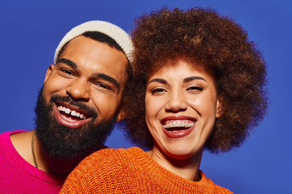Ένα ζευγάρι Αφροαμερικανών, ντυμένο με ζωντανή περιστασιακή ενδυμασία, χαμογελώντας ευτυχισμένοι μαζί σε ένα μπλε φόντο. - Φωτογραφία, εικόνα
