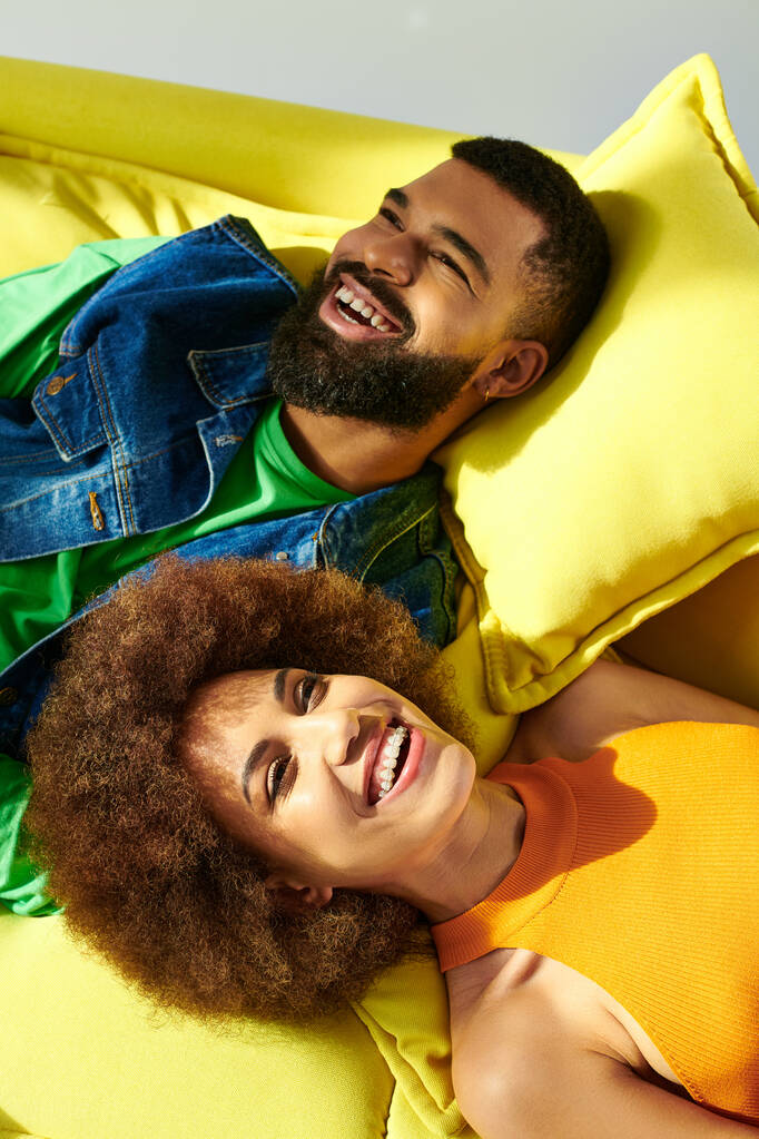 Canlı kıyafetler giymiş Afrikalı bir erkek ve kadın gri bir arka plana yaslanmış sarı bir yastığa uzanıyorlar.. - Fotoğraf, Görsel