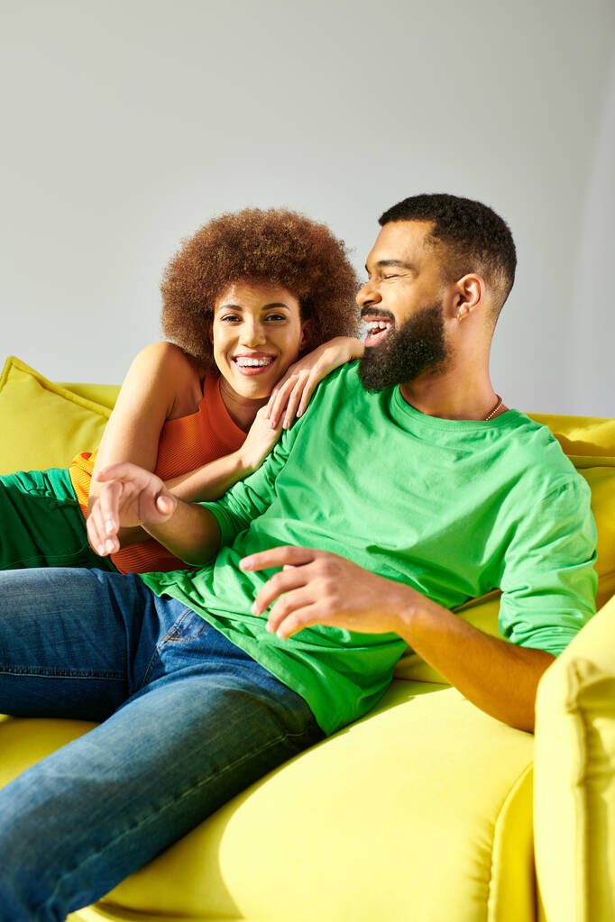 アフリカ系アメリカ人男性と女性は活気に満ちた服を着て,灰色の背景に黄色いソファに座っている間友情の瞬間を共有します.. - 写真・画像