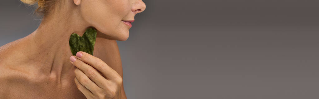 Alluring mature woman using gua sha on a gray backdrop. - Photo, Image