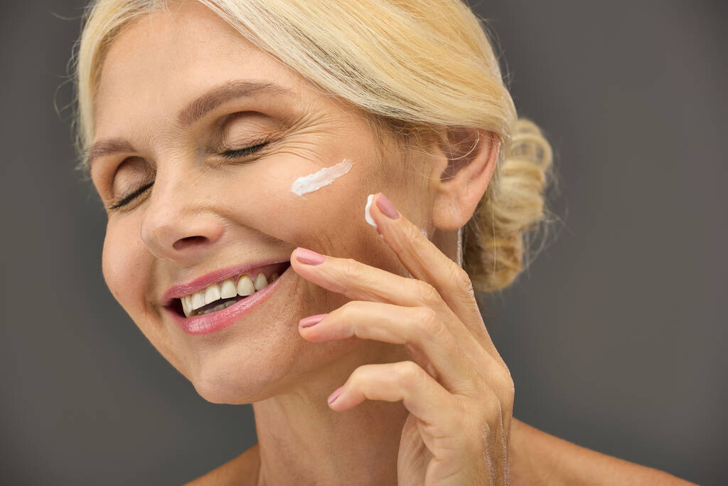Een volwassen vrouw brengt vrolijk huidverzorging crème op haar gezicht tegen een grijze achtergrond. - Foto, afbeelding