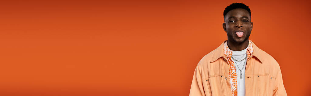 Молодой афроамериканец в стильной одежде высовывает язык на оранжевом фоне. - Фото, изображение