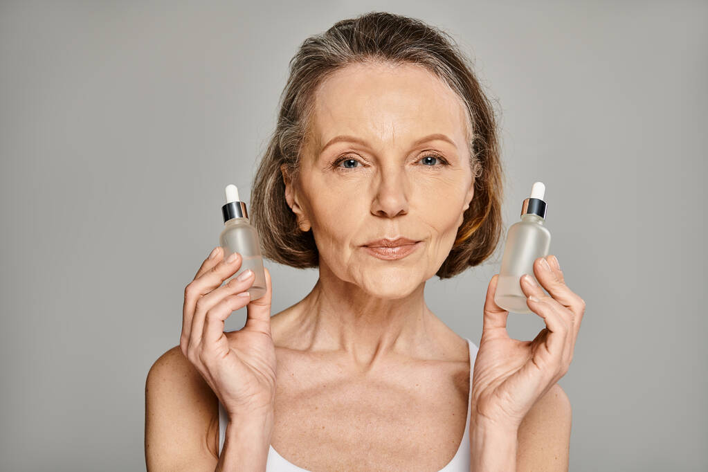 Una mujer madura y atractiva sostiene elegantemente dos botellas de productos para el cuidado de la piel. - Foto, Imagen