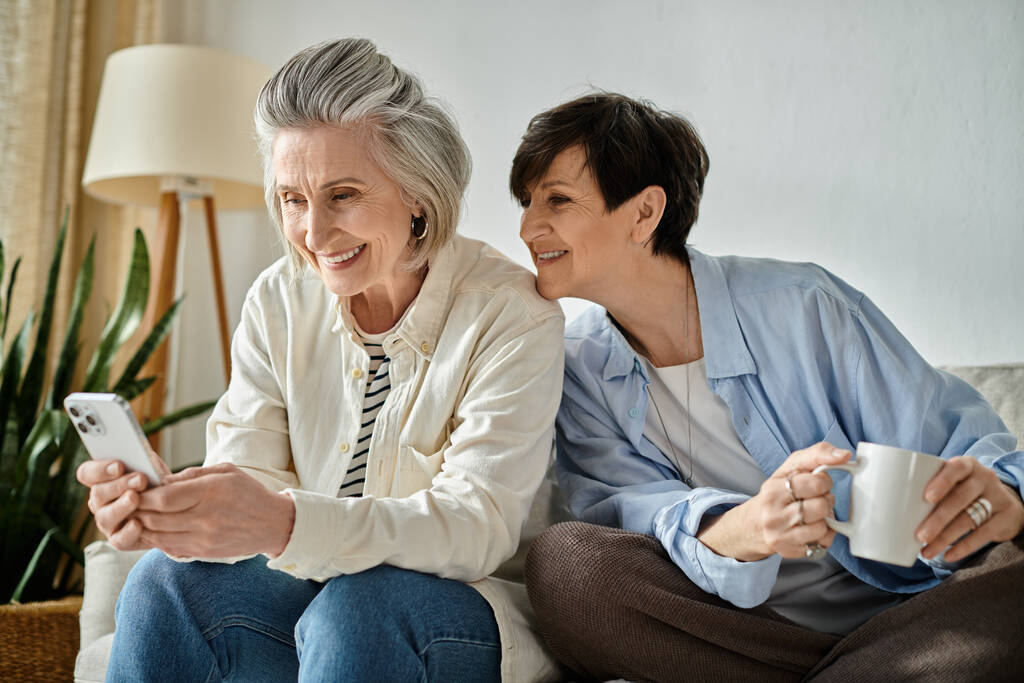 Zwei reife Frauen, ein liebendes lesbisches Paar, teilen einen Moment auf einer Couch, in ein Telefon vertieft. - Foto, Bild