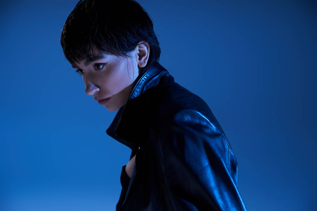 Trendy young woman in leather jacket poses confidently in front of a vivid blue background. - Foto, afbeelding