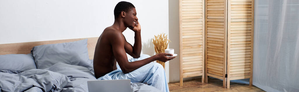 Mladý Afroameričan sedí na posteli, drží šálek kávy a užívá si klidné ranní chvíle. - Fotografie, Obrázek