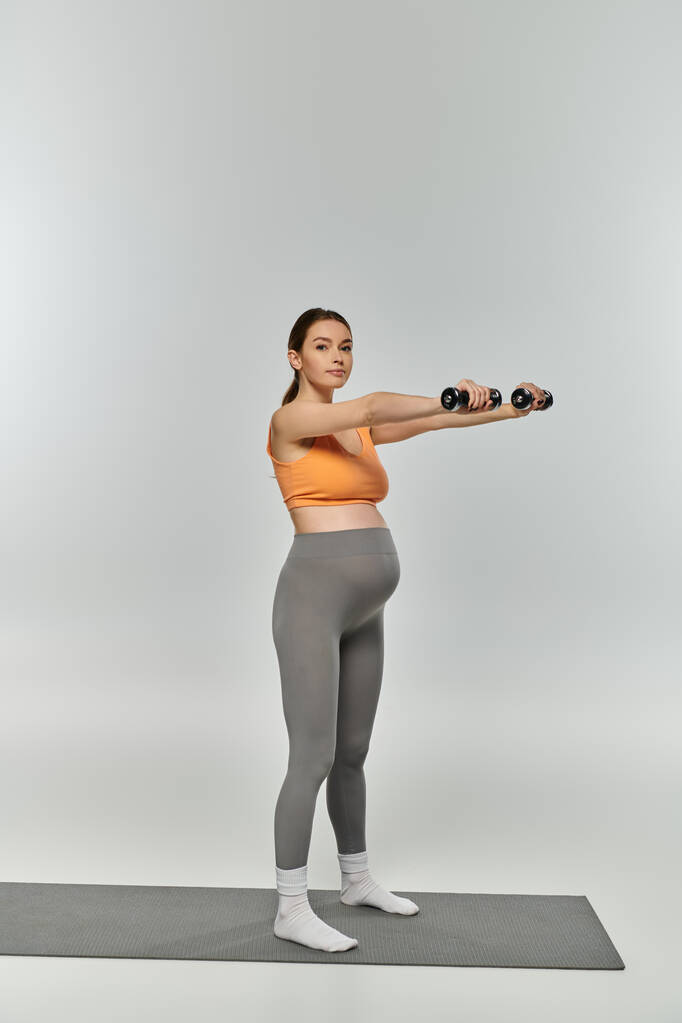 Una mujer embarazada deportiva en ropa deportiva realiza un ejercicio en cuclillas con una mancuerna sobre un fondo gris. - Foto, Imagen