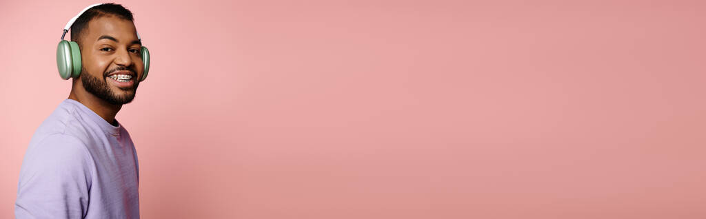 Яскравий молодий афроамериканський чоловік у фіолетовій сорочці та навушники посміхається на рожевому фоні. - Фото, зображення