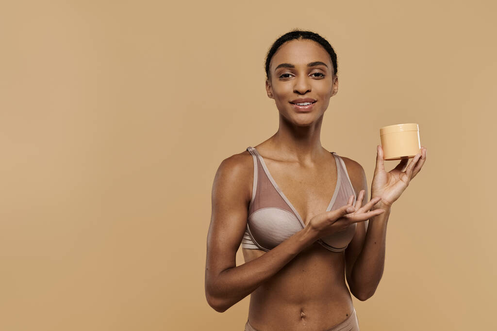 Afro-Amerikaanse vrouw, mooi en slank, staande in beha met body butter in de hand op een beige achtergrond. - Foto, afbeelding