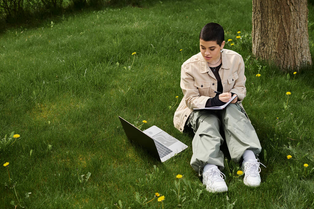 Молода жінка з коротким волоссям, в стильному повсякденному вбранні, сидить на зеленій траві на відкритому повітрі, працює на ноутбуці. - Фото, зображення