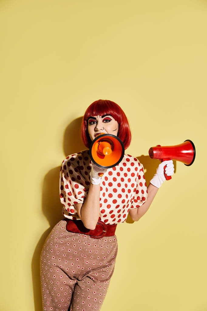 黄色の背景に対して赤とオレンジのメガフォンを保持するカラフルなメイクでポルカドットの赤毛の女性. - 写真・画像