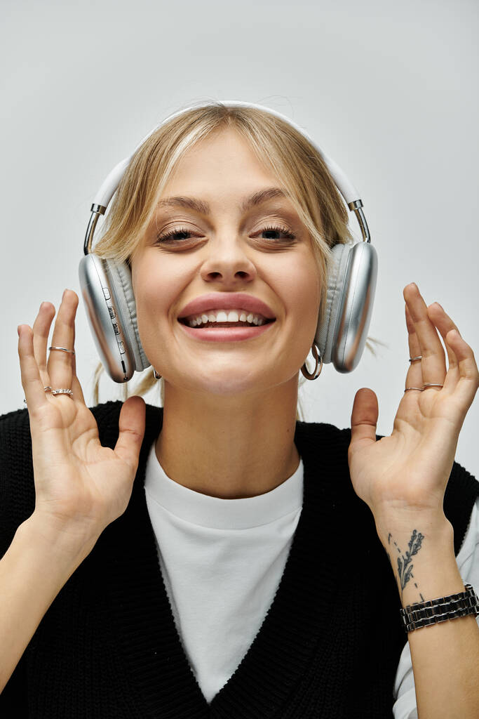 Μια νεαρή ξανθιά γυναίκα με μοντέρνα casual ενδυμασία χαμογελά ακούγοντας μουσική μέσω ακουστικών. - Φωτογραφία, εικόνα