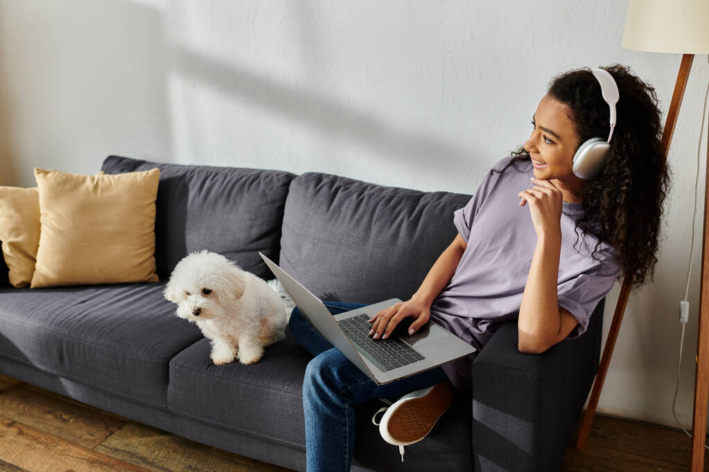 Mujer sentada en el sofá con el ordenador portátil, acariciando su perro bichon frise. - Foto, imagen