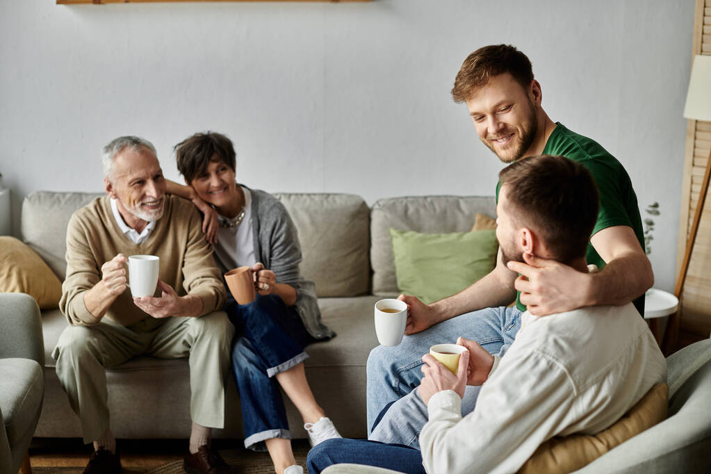 Una pareja gay se sienta en un sofá, abrazado y sosteniendo copas, con los padres en el fondo. - Foto, Imagen
