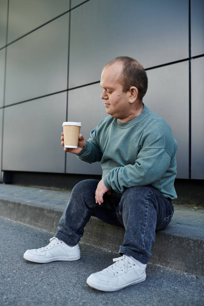 Mies, jolla on osallisuutta, istuu jalkakäytävällä pitämässä kahvikuppia, joka näyttää olevan ajatuksissaan.. - Valokuva, kuva
