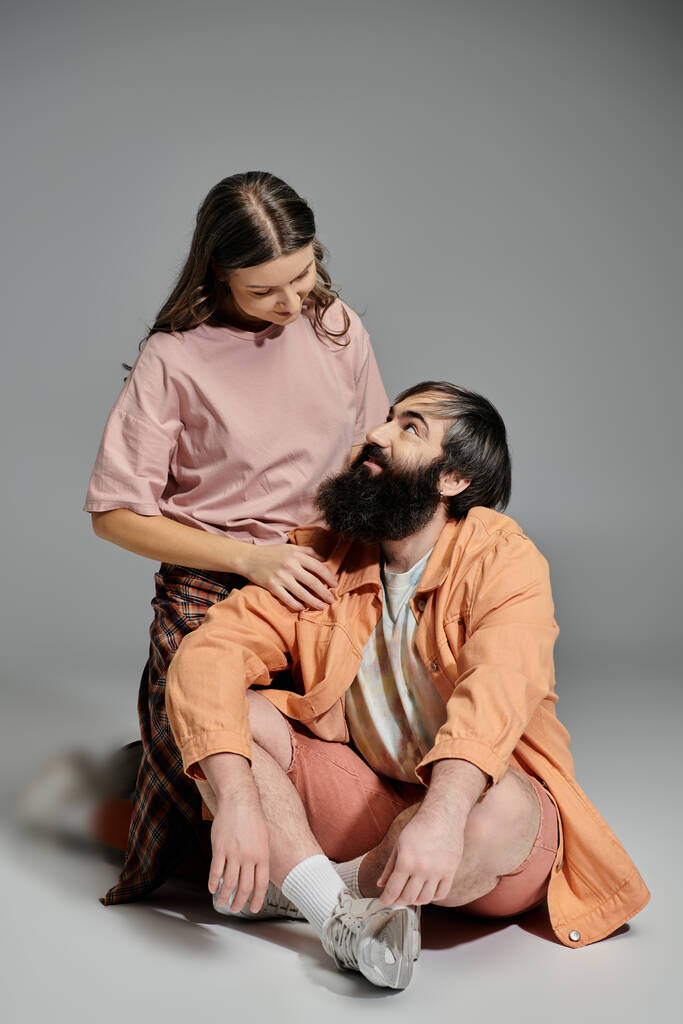 Um par senta-se em um cenário de estúdio, a mulher em uma camisa rosa e calças xadrez, o homem em um casaco laranja e shorts, olhando um para o outro com amor. - Foto, Imagem