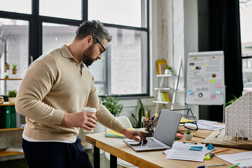 Un hombre de negocios guapo con barba trabaja en su portátil en un espacio de oficina moderno, sosteniendo una taza de café en una mano. - Foto, Imagen