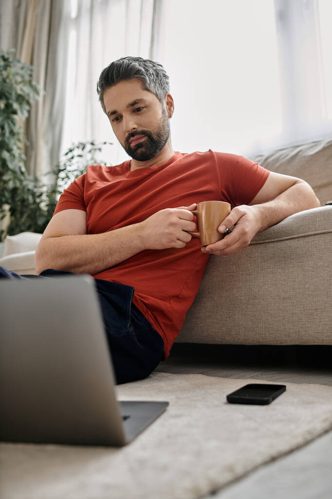 Un hombre barbudo con atuendo casual se sienta en el suelo, trabajando remotamente en su computadora portátil con una taza de café en la mano. - Foto, imagen
